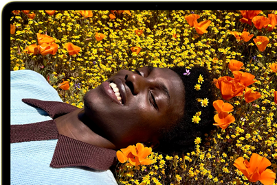 MacBook Neo, citrus color, screen shows a smiling person lying in a sunny field of flowers